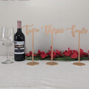 May include: A wine glass, a bottle of red wine, and three wooden table number signs that read "Two Three Four" are arranged on a white tablecloth. Red flowers and greenery are at the base of the signs.