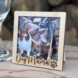 Puede incluir: Un marco de fotos de madera con una foto de dos perros. El marco tiene las palabras "Dog Mama" y una huella de pata. Los perros son una mezcla de marrón, negro y blanco.