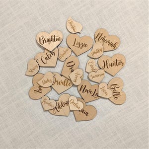 May include: A collection of wooden heart-shaped cutouts with engraved names in a variety of fonts. The names include "Brighton", "Lizzie", "Hunter", and others. The cutouts are scattered on a light-colored fabric.