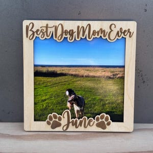 Puede incluir: Un marco de fotos de madera con las palabras "Best Dog Mom Ever" y "June" grabadas. El marco contiene una foto de un perro en un campo de hierba bajo un cielo azul. Huellas de patas también están grabadas en el marco.