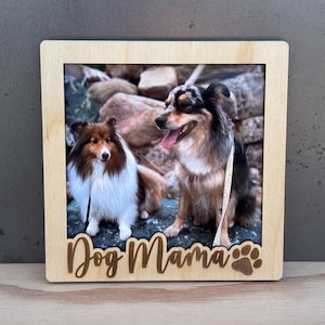 Puede incluir: Un marco de fotos de madera con una foto de dos perros. El marco tiene las palabras "Dog Mama" y una huella de pata tallada en la madera. Los perros son una mezcla de marrón, negro y blanco, con la lengua de un perro fuera.