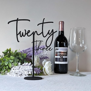 May include: A black metal table number stand with the words "Twenty Three" in a cursive font. A bottle of red wine, a wine glass, and floral arrangement with white and purple flowers are next to the stand.