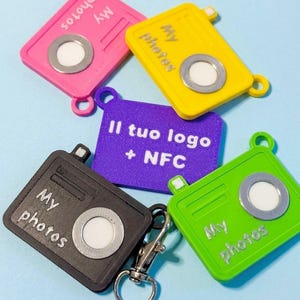 Può includere: Una collezione di portachiavi quadrati colorati, rosa, giallo, verde, nero e viola. Ogni portachiavi presenta un design a fotocamera con le parole "My photos" o "Il tuo logo + NFC". Un fermaglio argentato è attaccato al portachiavi nero.
