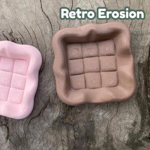 Puede incluir: Dos bandejas cuadradas con bordes ondulados. Una es rosa claro, la otra marrón. Cada una tiene un patrón de cuadrícula de nueve cuadrados. El texto "Retro Erosion" está en la esquina superior derecha.