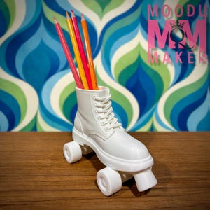 May include: A white roller skate-shaped pencil holder with colorful pencils. The pencil holder is a white boot design with laces and wheels. The background is a retro blue, green, and white pattern. The text "MOODY MOM MAKES" is in pink.