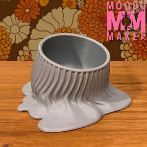 May include: A gray, 3D-printed planter with a unique, melted design. The planter has a cylindrical opening and a base that appears to be flowing or melting. The text "MOODY MOM MAKES" is visible in the background.