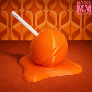 May include: An orange, spherical pen holder with a white stylus inserted. The pen holder sits on a matching orange, melted-looking base. The background features a retro orange and brown patterned wallpaper. The text "MOODY MOM MAKES" is in the upper right corner.