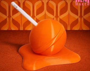 May include: An orange, spherical pen holder with a white stylus inserted. The pen holder sits on a matching orange, melted-looking base. The background features a retro orange and brown patterned wallpaper. The text "MOODY MOM MAKES" is in the upper right corner.