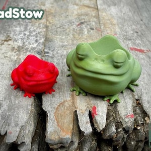 May include: Two frog-shaped planters, one red and one green, are sitting on a wooden surface. The green frog planter has a wide mouth, perfect for planting small plants.