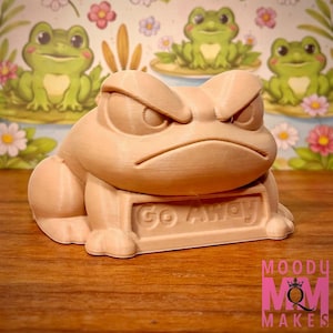 May include: A light brown, 3D-printed frog figurine with an angry expression, holding a sign that reads "Go Away." The background features a floral pattern with cartoon frogs. The figurine is a unique decorative item.