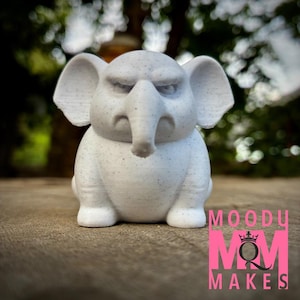 May include: A small, light gray elephant figurine with a grumpy expression. The elephant has large ears, a trunk, and visible speckles. The figurine is sitting on a wooden surface. The text "MOODY MOM MAKES" is in pink.
