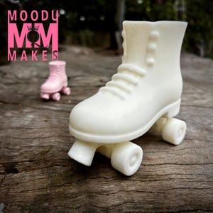 Puede incluir: Una figura de patín de ruedas de color crema con cordones y ruedas visibles. Un patín de ruedas rosa más pequeño está en el fondo. El logotipo "MOODY MAKES" está en rosa en la parte superior izquierda. Sobre una superficie de madera envejecida.