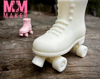 May include: A large, off-white roller skate figurine with visible laces and wheels. A smaller, pink roller skate is in the background. The logo "MOODY MAKES" is in pink at the top left. The items are on a weathered wooden surface.