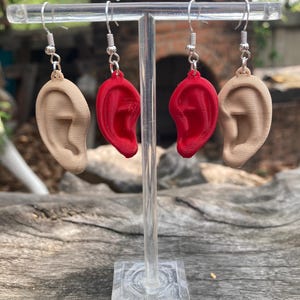 May include: A pair of earrings featuring a 3D printed ear design. One earring is red and the other is a skin tone color. The earrings are displayed on a clear acrylic stand.