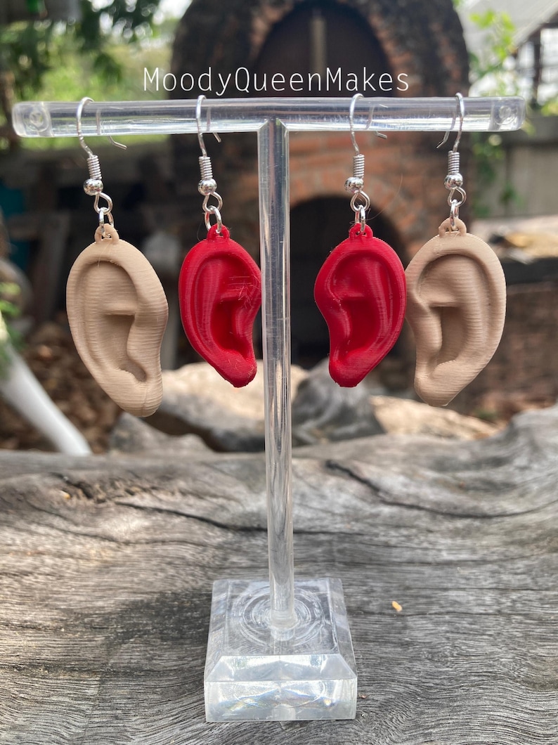 May include: A pair of earrings featuring a 3D printed ear design. One earring is red and the other is a skin tone color. The earrings are displayed on a clear acrylic stand.