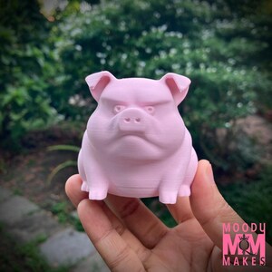 May include: A light pink, 3D-printed pig figurine with an angry expression. The pig has a furrowed brow, small ears, and a stout body. The figurine is held in a hand against a blurred green background.