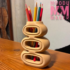 May include: A light brown, 3-tiered pencil holder with oval cutouts, holding several coloured pencils. The pencils are red, pink, orange, yellow, green, and blue. The text "MOODY MOM MAKES" is in pink.