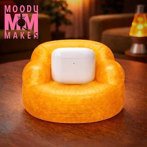 May include: A white wireless earbud charging case rests on a small, bright orange, miniature armchair. The chair is on a dark wooden surface. The text "MOODU MOM MAKES" is in pink letters in the upper left corner.