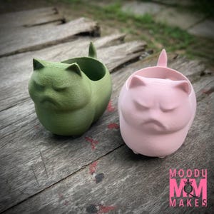 Puede incluir: Dos macetas con forma de gato en verde y rosa, cada una con una expresión de mal humor y una pequeña cola. Las macetas están sobre una superficie de madera desgastada. El logotipo "MOODY MOM MAKES" es visible en la esquina inferior derecha.