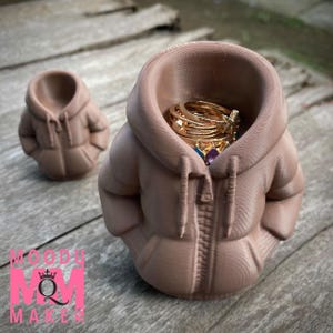 Op de afbeelding: Een lichtbruine, 3D-geprinte ringhouder in de vorm van een hoodie. De hoodie heeft een rits en capuchon, met een kleinere versie op de achtergrond. De grotere hoodie bevat meerdere gouden ringen met edelstenen. Het logo "MOODY MOM MAKES" is zichtbaar.