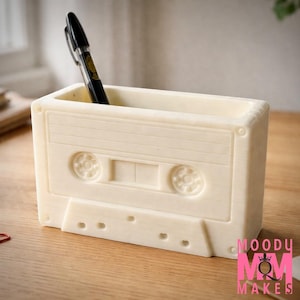 May include: Off-white cassette tape-shaped desk organizer with a black pen inside. The organizer has a rectangular shape with detailed molding, including the tape reels and other features of a cassette. The organizer is on a wooden surface.