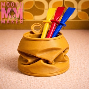 May include: A mustard-coloured, crushed can-shaped container holding yellow, red, and blue spatulas. The container has a textured surface and the text "MOODY MOM MAKES" in pink.