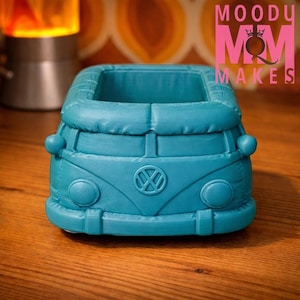 May include: A teal-colored planter shaped like a vintage Volkswagen bus. The planter has rounded headlights, a VW logo, and a rectangular opening for plants. The text "MOODY MOM MAKES" is visible in pink.