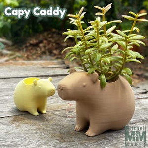 May include: Two 3D printed planter pots shaped like capybaras. The larger pot is brown and holds a small green plant. The smaller pot is yellow. The text "Capy Caddy" is visible in the top left corner of the image.