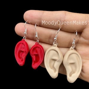May include: A pair of earrings featuring a 3D printed ear design. One pair is red and the other is a light beige color. The earrings are on silver hooks.