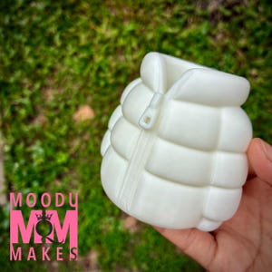 May include: A white, puffy vest-shaped object with a zipper is held in a hand against a blurred green background. The vest is a solid, light color and has a quilted design. The text "MOODY MOM MAKES" is visible in pink.
