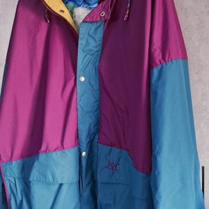 Könnte beinhalten: Farbblock-Jacke in Lila, Türkis und Gelb. Die Jacke hat eine Kapuze, Taschen und einen Kordelzug in der Taille. Das Logo "HH" ist auf dem türkisfarbenen Bereich gestickt. Die Jacke hängt an einem schwarzen Kleiderbügel.