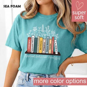 Puede incluir: Camiseta verde espuma de mar con el texto "Applied Behavior Analysis". La camiseta presenta un gráfico de libros con títulos como "Prompt" y "Fade". También se ven las palabras "super soft" y "more color options".