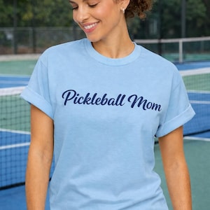 Può includere: T-shirt azzurra con maniche arrotolate con la scritta "Pickleball Mom" in corsivo blu scuro. La persona sorridente indossa la maglietta davanti a un campo da tennis.