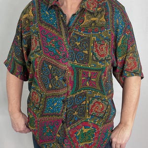 May include: A button-down shirt with a mosaic-like pattern in shades of green, blue, pink, and yellow. The short-sleeved shirt has a collared neckline and a pocket on the left chest. The shirt is worn with blue jeans.