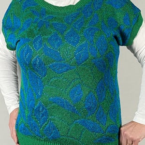 May include: A green and blue leaf-patterned sweater. The short-sleeved sweater has a crew neck and a green ribbed hem. The leaf pattern is in shades of blue and green. A white long-sleeved shirt is worn underneath.