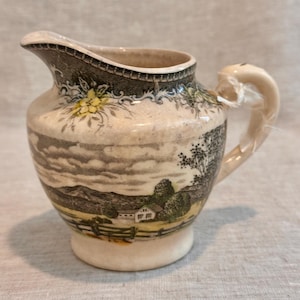 May include: A cream-colored ceramic creamer featuring a transferware landscape scene with a house, trees, and mountains. The rim is decorated with a dark gray floral pattern and yellow accents. It has a curved handle and a pouring spout.