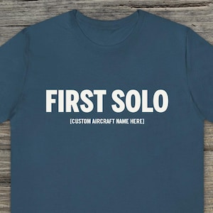 Puede incluir: Una camiseta azul marino con las palabras "FIRST SOLO" en letras blancas en negrita. Debajo del texto, en letras blancas más pequeñas, se lee "[CUSTOM AIRCRAFT NAME HERE]" en la parte delantera de la camiseta. La camiseta se muestra sobre un fondo de madera.