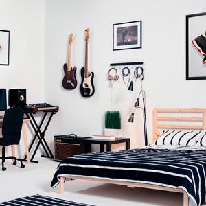K&ouml;nnte beinhalten: Ein Schlafzimmer mit einem Bett, einem Schreibtisch und einer Gitarre, die an der Wand h&auml;ngt. Das Bett hat eine schwarz-wei&szlig; gestreifte Bettdecke. Der Schreibtisch hat einen Computer, eine Tastatur und zwei Lautsprecher. Die Gitarre ist schwarz und hat ein wei&szlig;es Schlagbrett. An der Wand h&auml;ngt ein gerahmtes Bild von einem Paar rot-schwarzer Turnschuhe.