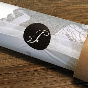 K&ouml;nnte beinhalten: Eine wei&szlig;e Posterrolle mit einem schwarzen Kreis, der einen wei&szlig;en Buchstaben F enth&auml;lt. Die Rolle ist in wei&szlig;es Seidenpapier eingewickelt und hat eine braune Pappkappe.