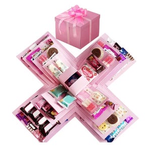 以下が含まれることがあります： ピンク色の爆発ボックス。ピンクのリボンで結ばれたお揃いの蓋が付いています。箱は開いており、個別に包装されたキャンディー、チョコレート、お菓子が詰められた複数のコンパートメントが現れています。