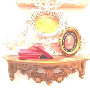Puede incluir: Un estante de pared de madera con tallas ornamentadas sostiene una pila de libros rojos y un peque&ntilde;o marco ovalado que contiene un retrato. Detr&aacute;s del estante, un gran marco ovalado decorativo pintado de blanco con un centro amarillo.