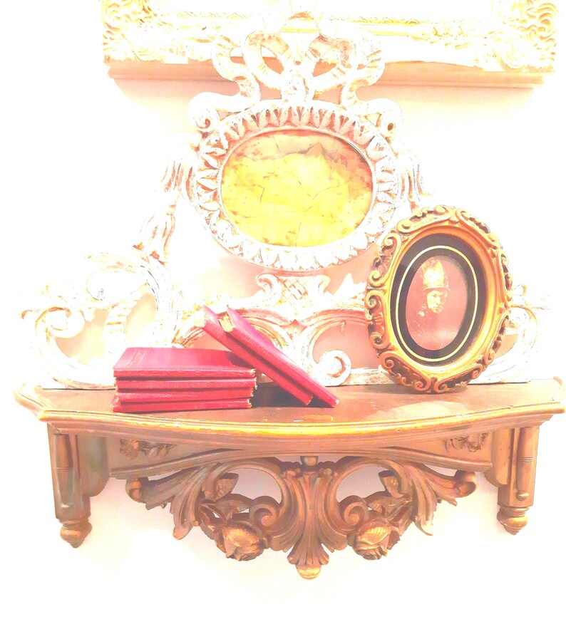 Puede incluir: Un estante de pared de madera con tallas ornamentadas sostiene una pila de libros rojos y un peque&ntilde;o marco ovalado que contiene un retrato. Detr&aacute;s del estante, un gran marco ovalado decorativo pintado de blanco con un centro amarillo.