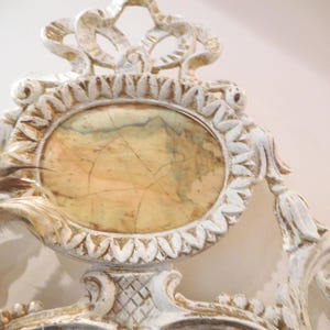 Puede incluir: Un marco ornamentado blanco y dorado con un inserto ovalado central de patr&oacute;n jaspeado en beige y marr&oacute;n. El marco presenta detalles tallados intrincados y volutas, con una pluma marr&oacute;n y blanca sujeta en el lado izquierdo.