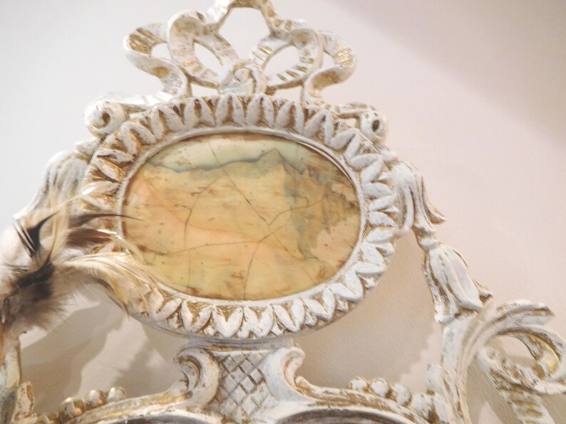 Puede incluir: Un marco ornamentado blanco y dorado con un inserto ovalado central de patr&oacute;n jaspeado en beige y marr&oacute;n. El marco presenta detalles tallados intrincados y volutas, con una pluma marr&oacute;n y blanca sujeta en el lado izquierdo.