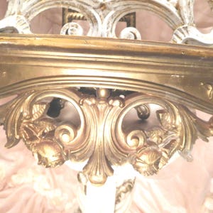 Puede incluir: Un estante de pared con acabado en tono dorado, que presenta intrincados tallados de volutas y motivos florales. El estante tiene una superficie superior horizontal lisa y un soporte decorativo ornamentado debajo con relieves detallados.