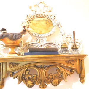 Puede incluir: Un estante de pared dorado con tallas florales ornamentadas y un panel trasero decorativo a juego con un centro ovalado. En el estante hay una figura de p&aacute;jaro sobre un pedestal, dos libros apilados y dos tinteros de vidrio transparente.