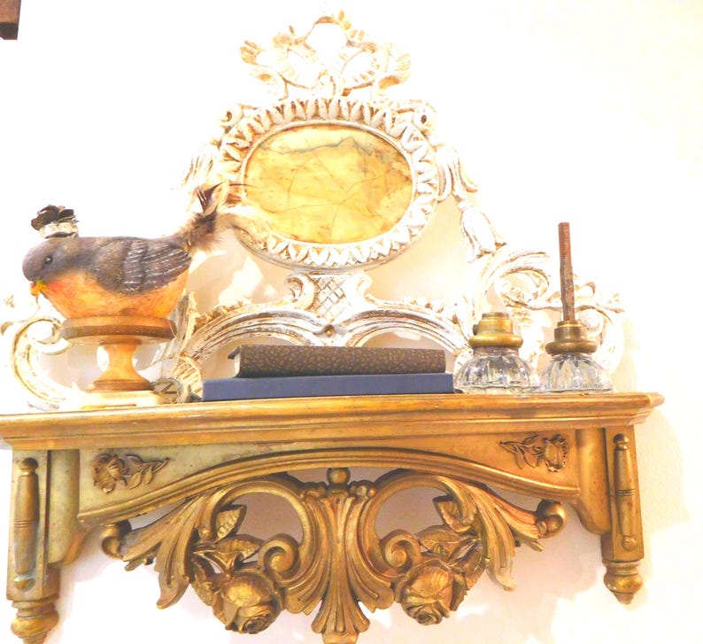 Puede incluir: Un estante de pared dorado con tallas florales ornamentadas y un panel trasero decorativo a juego con un centro ovalado. En el estante hay una figura de p&aacute;jaro sobre un pedestal, dos libros apilados y dos tinteros de vidrio transparente.