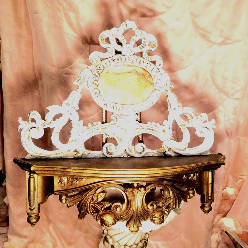 Puede incluir: Una consola de pared con base dorada que presenta tallas ornamentales de volutas y detalles florales. Sobre el estante se sit&uacute;a un gran remate decorativo blanco con un panel central ovalado de acabado jaspeado en amarillo claro.