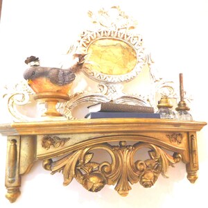 Puede incluir: Un estante de pared dorado con tallas florales y volutas ornamentadas. Sobre el estante hay una peque&ntilde;a figura de p&aacute;jaro con plumas marrones y naranjas, dos libros oscuros apilados y dos tinteros de vidrio transparente.
