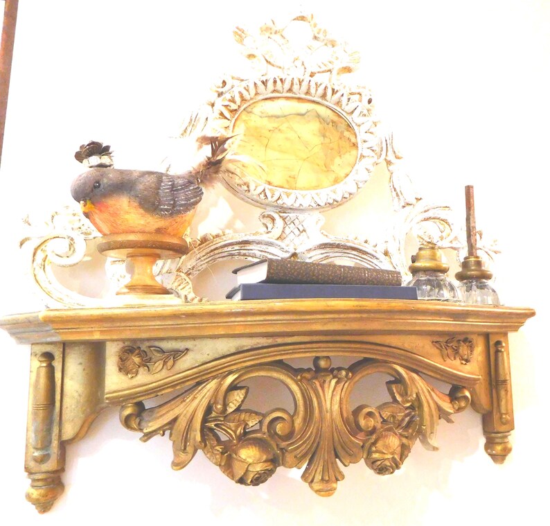 Puede incluir: Un estante de pared dorado con tallas florales y volutas ornamentadas. Sobre el estante hay una peque&ntilde;a figura de p&aacute;jaro con plumas marrones y naranjas, dos libros oscuros apilados y dos tinteros de vidrio transparente.